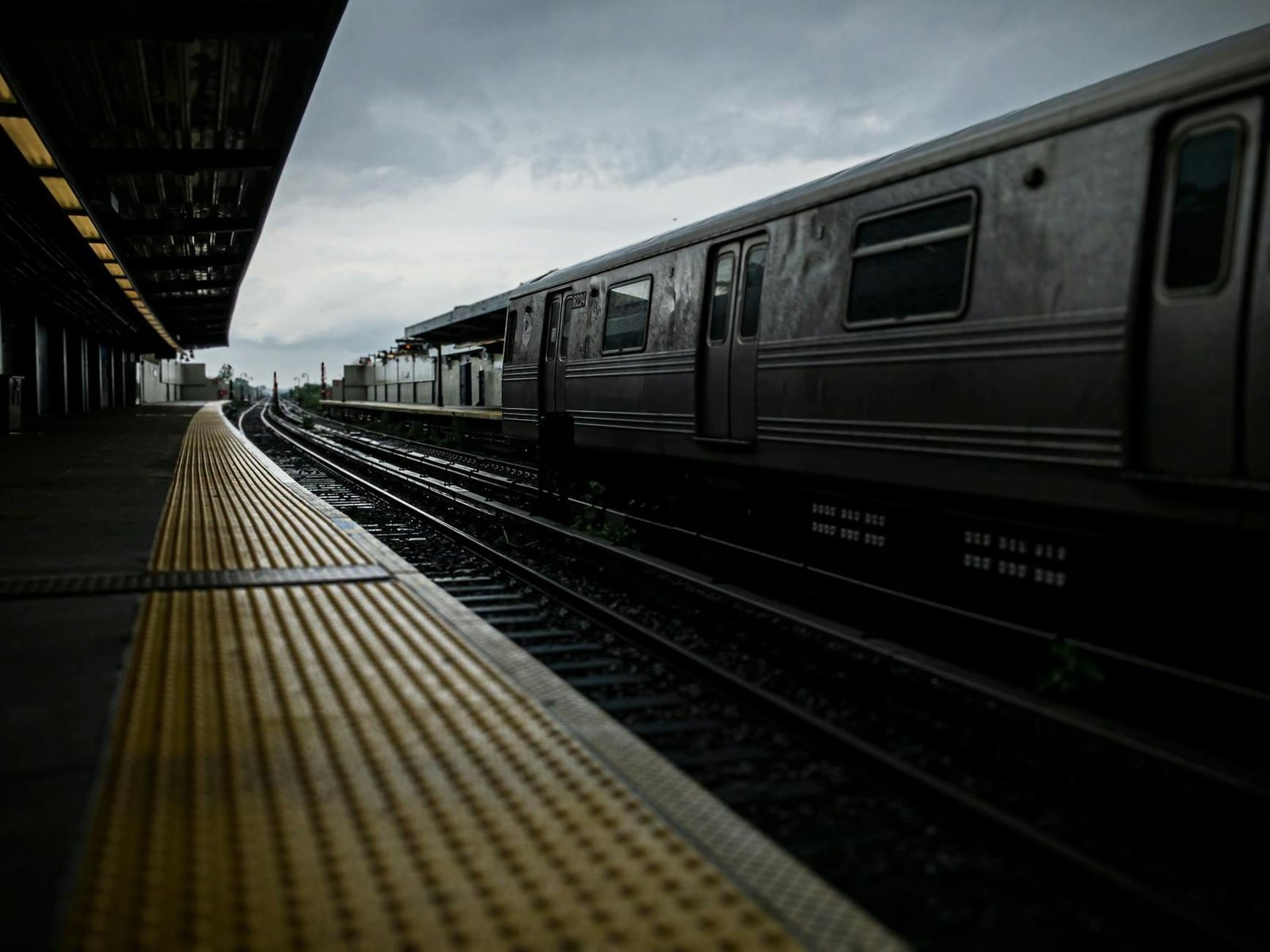 Estación de metro elevada de Nueva York con tren gris, vías y andén amarillo bajo cielo nublado.