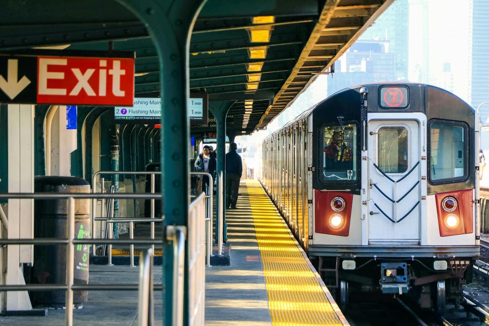 NYC 7 train at an elevated subway station platform with an exit sign and sunlight.