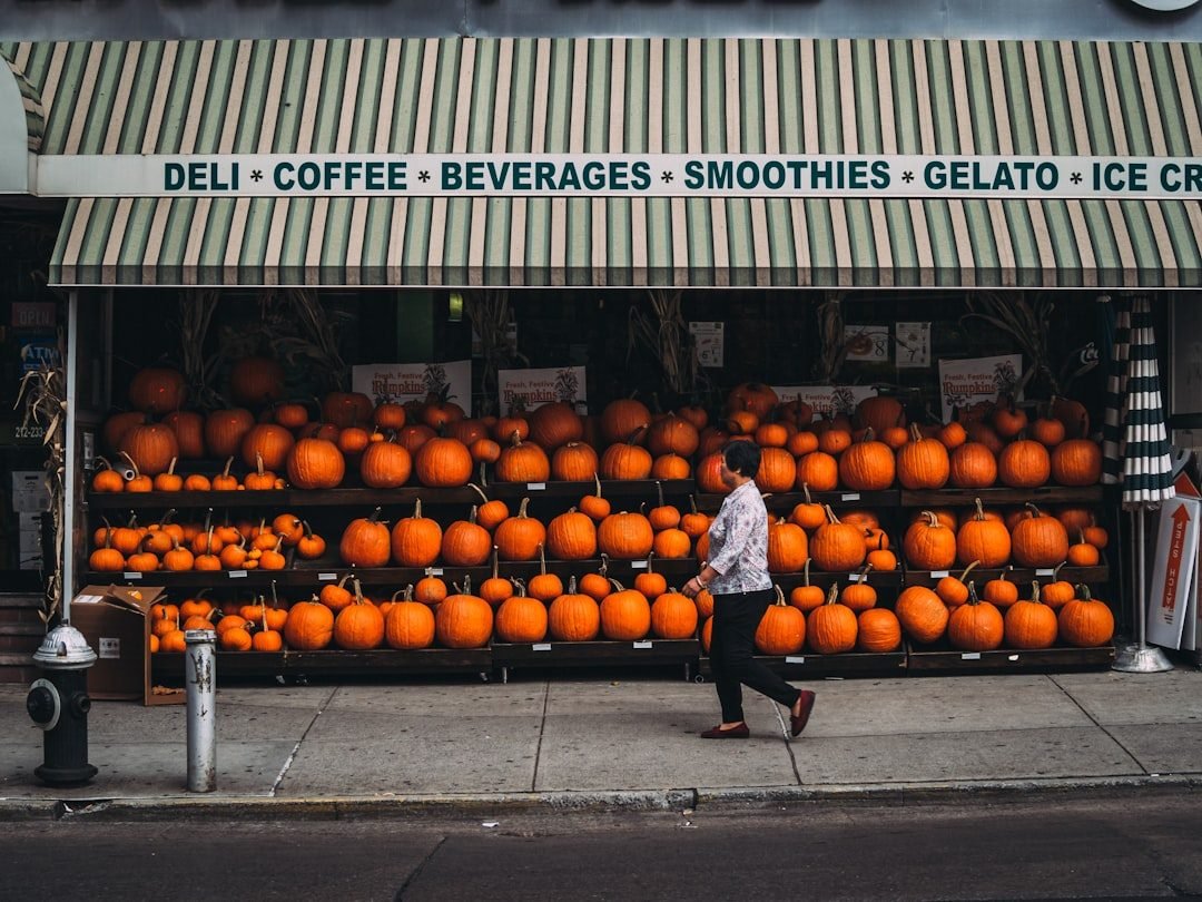NYC deli storefront with fall pumpkin display and striped awning, a woman walks by.
