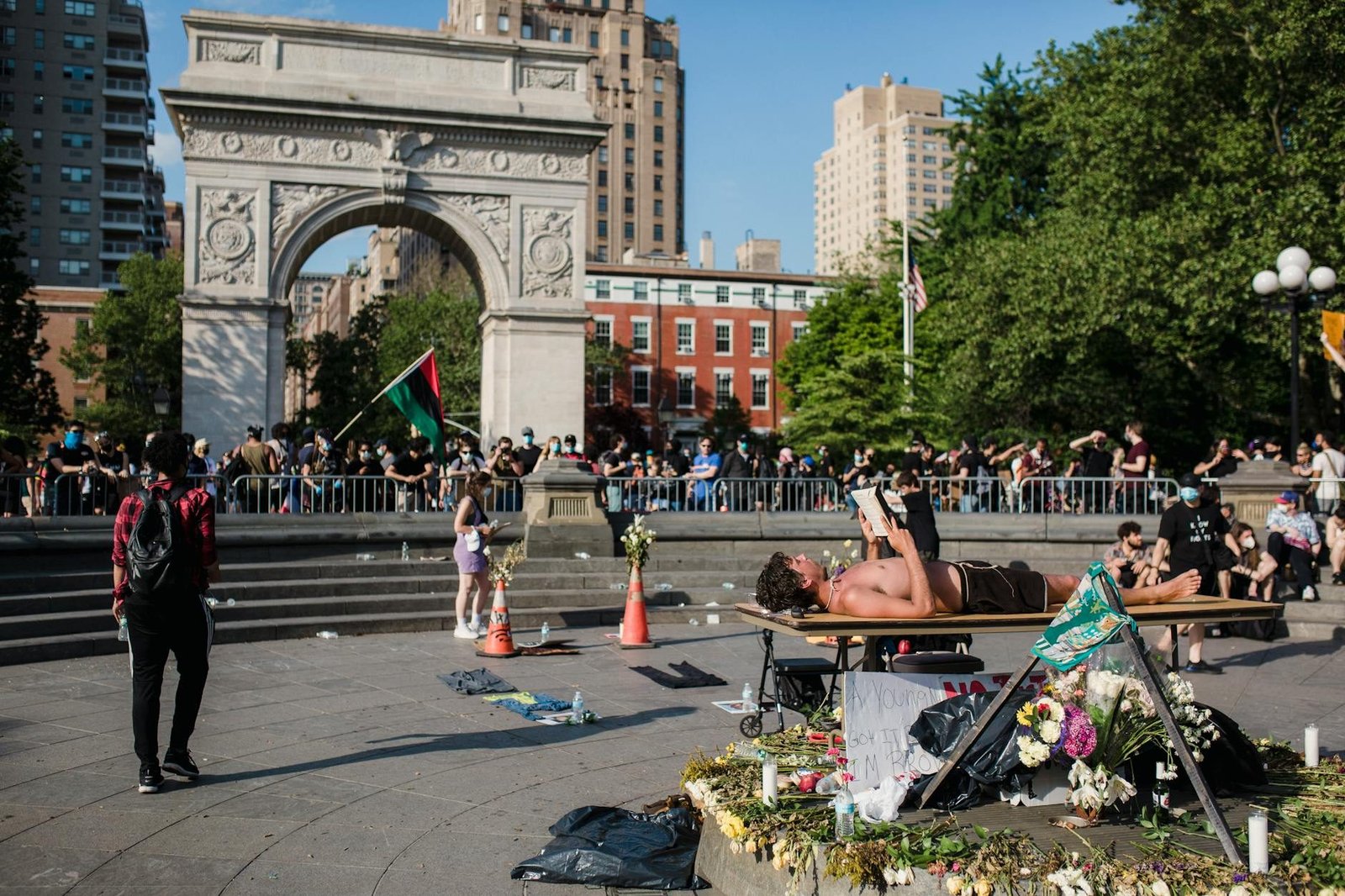 Washington Square Park Arch, sunny day, man reading, flowers, and crowd gathering.